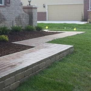 A brick walkway with a low brick retaining wall on one side winds across a green lawn toward a garage door.