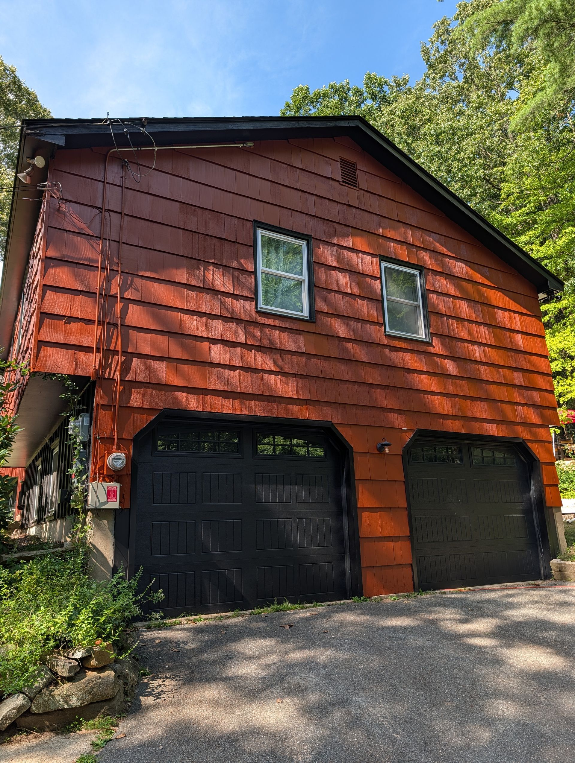 Red wooden garage with black doors on a gravel driveway, surrounded by trees and greenery.