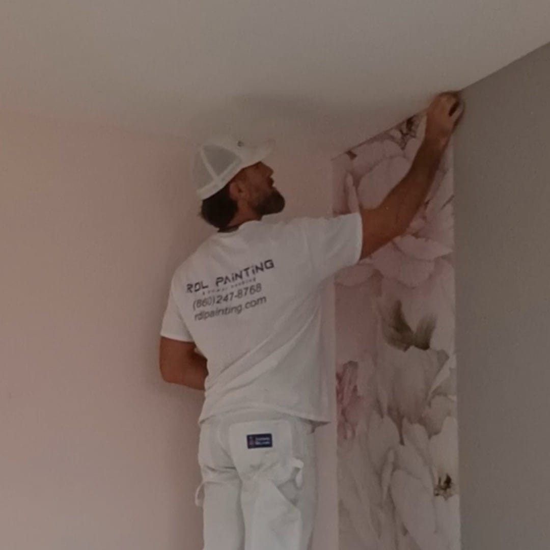 Painter in white uniform hangs floral wallpaper on a wall corner.