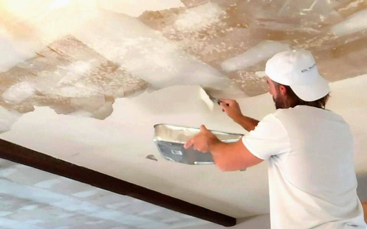 Worker plastering a ceiling with a trowel and mud pan in a white room