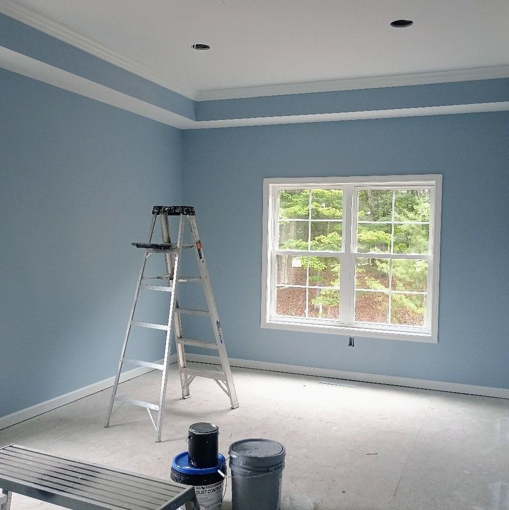 Blue empty room being painted, with ladder, paint bucket, and a sunlit window