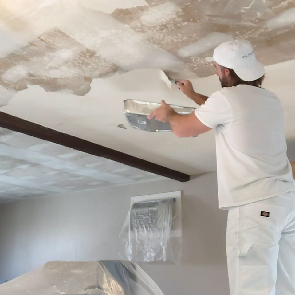 Worker in white repainting a ceiling with a putty knife and tray in a room under renovation