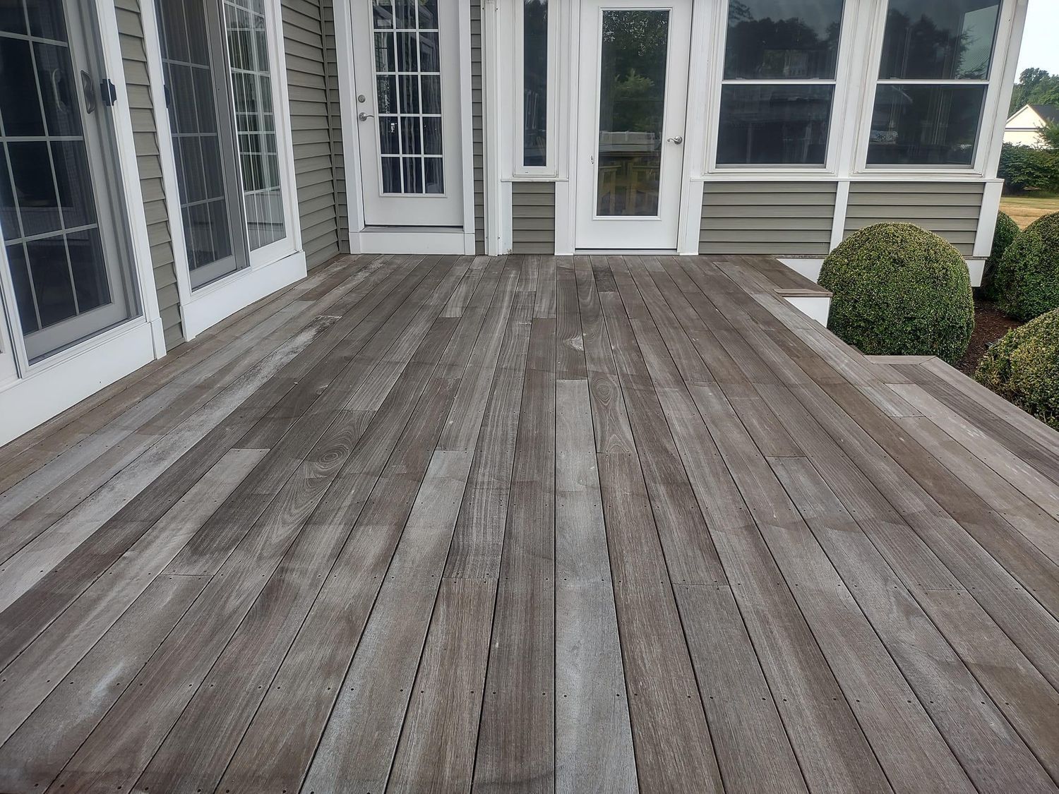 Weathered gray wooden deck leading to a house door, with windows on the left and shrubs on the right.