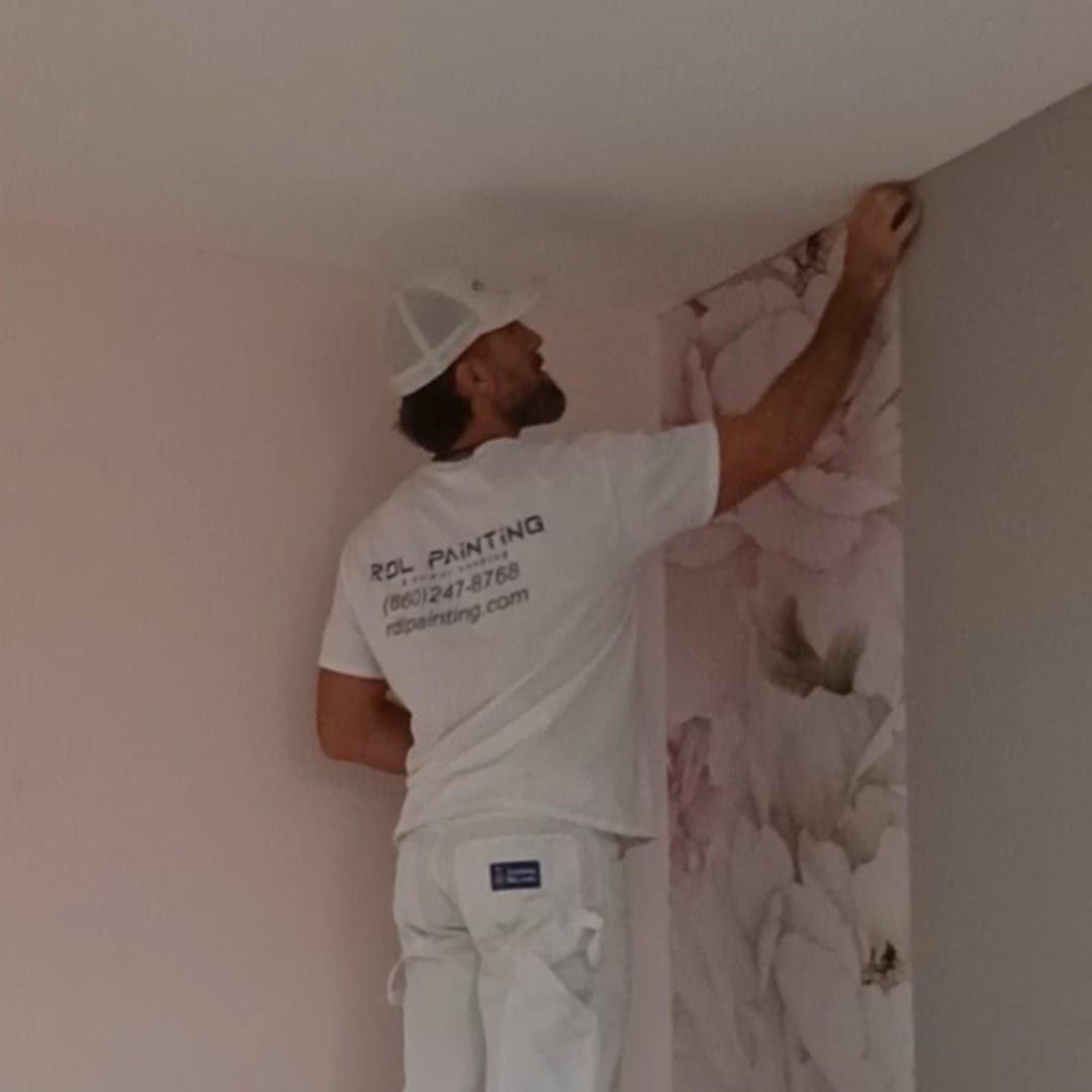 Man hanging pink floral wallpaper in a corner of a room