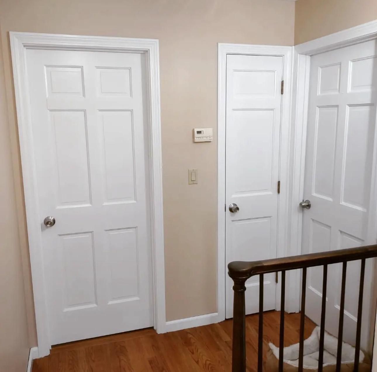 Hallway with two white doors, a thermostat, and a staircase railing on hardwood floors.