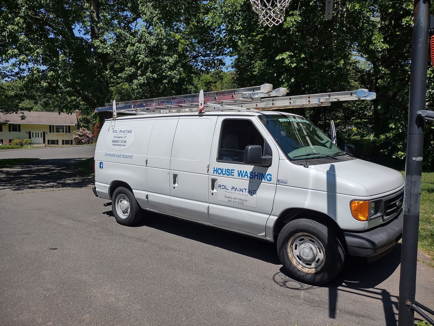 White utility van parked on a street, with ladders on top and trees in the background.