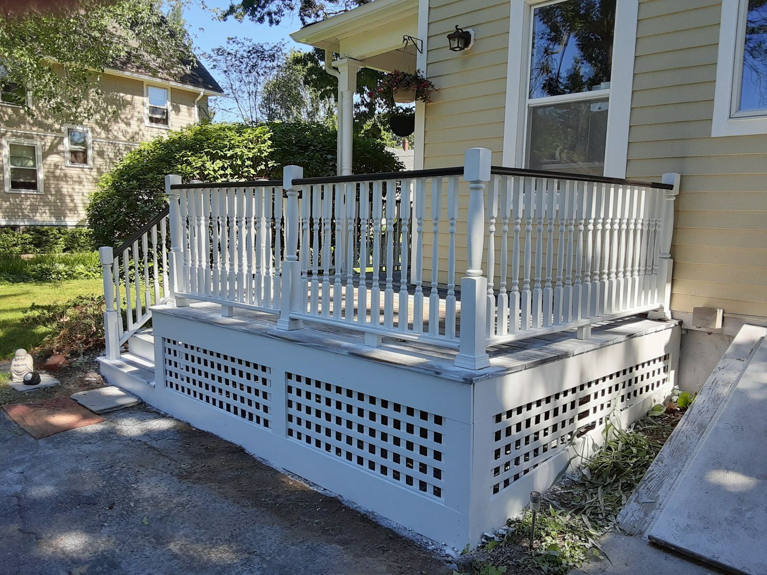 White-railed porch with lattice skirting on a beige house, beside a driveway and trees