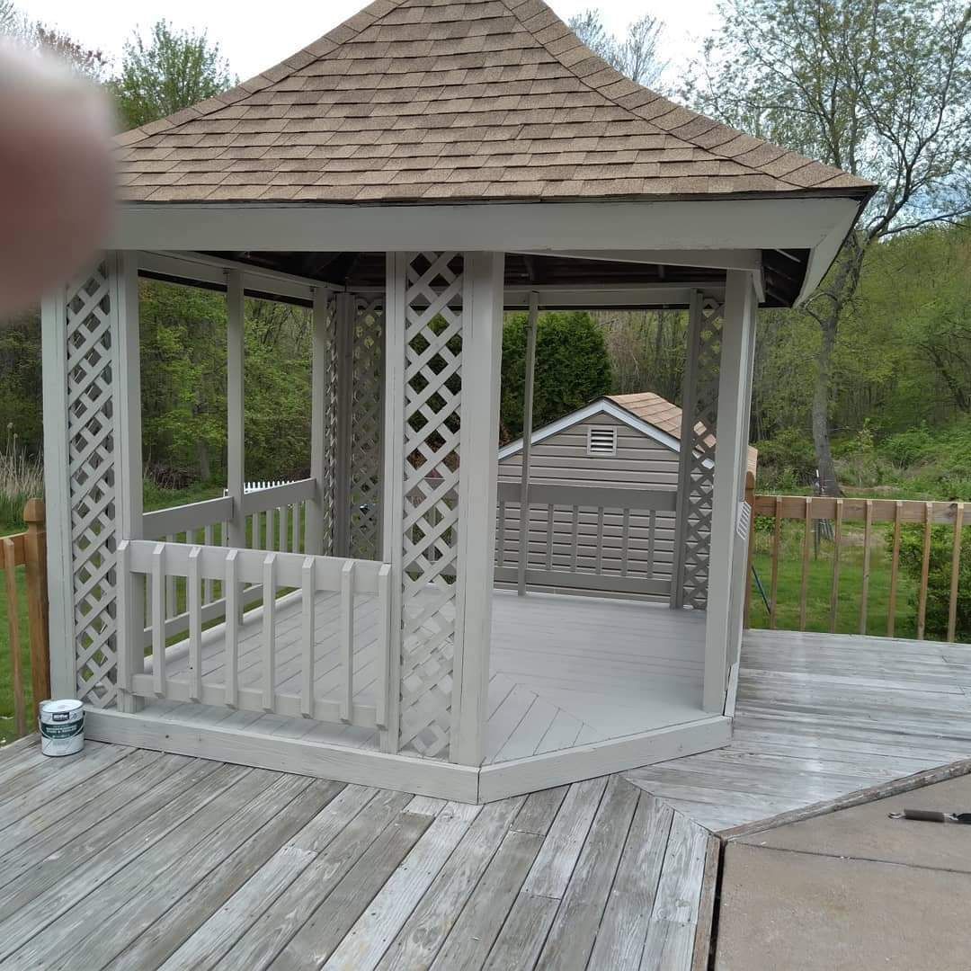 Wooden gazebo with lattice sides on a deck, viewed outdoors with trees and a fingertip in the upper left.