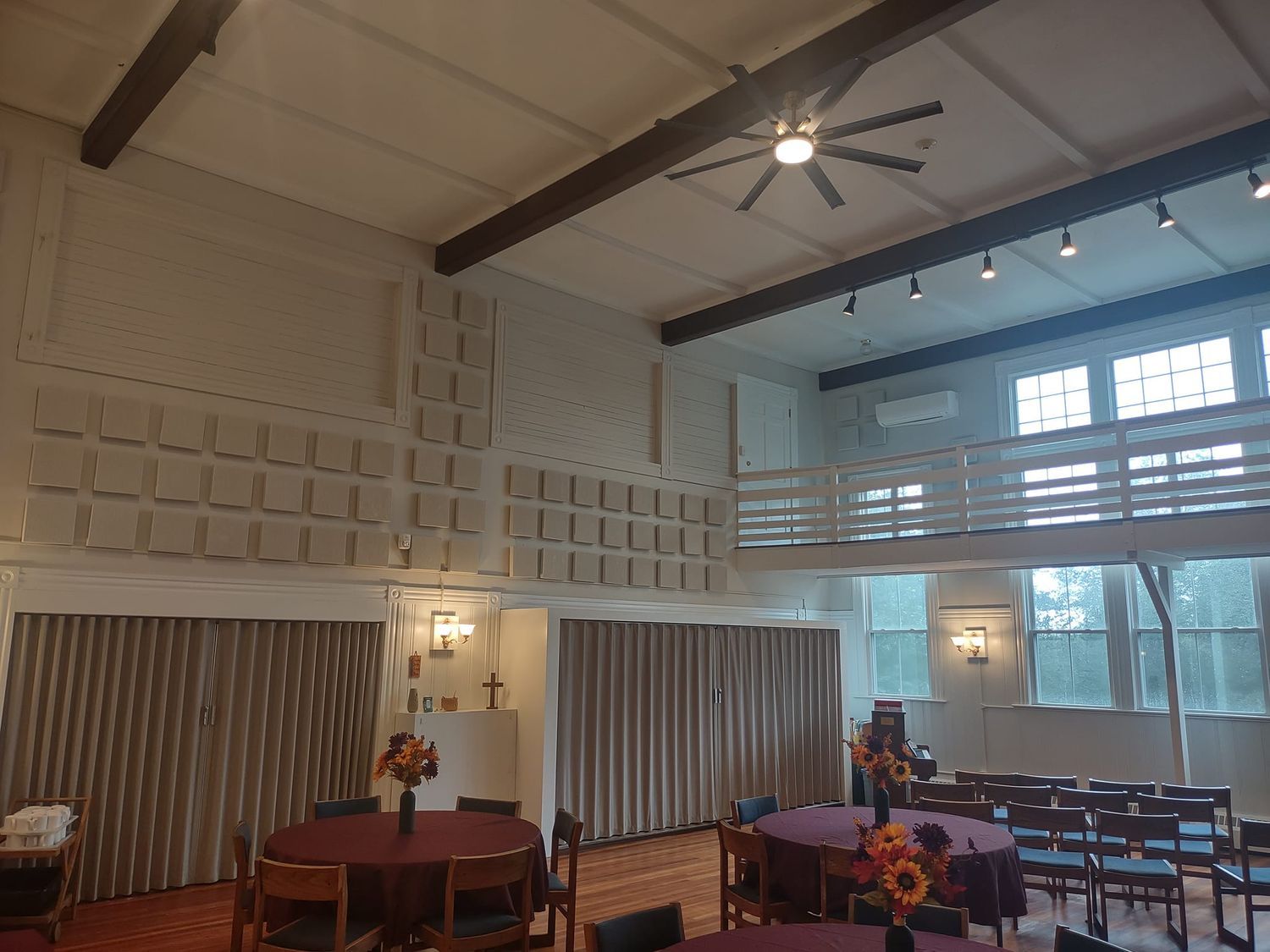 Dining hall with round tables, striped curtains, and tall windows under a vaulted ceiling with a ceiling fan.