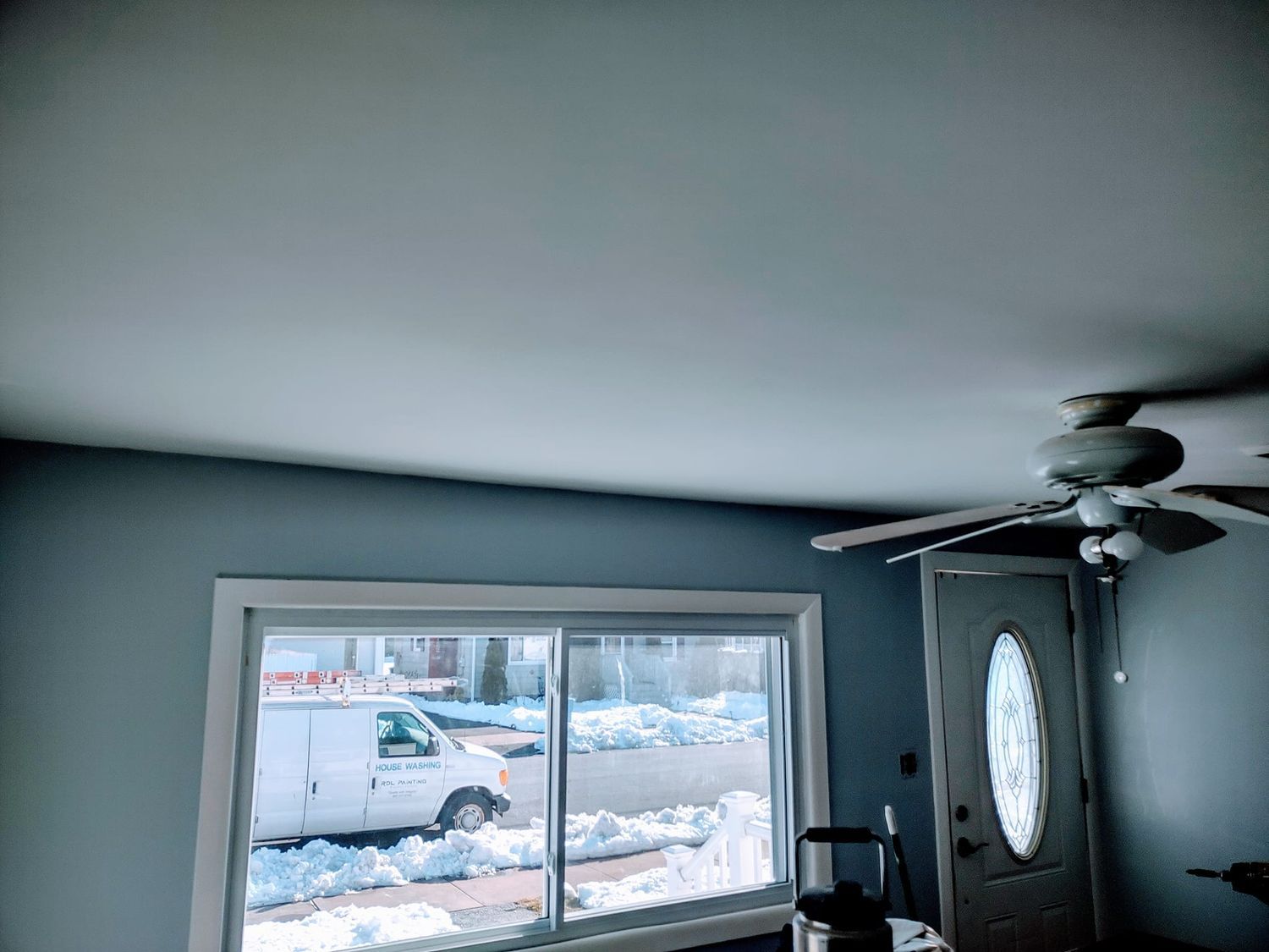 Snowy street viewed through a window in a gray living room with a ceiling fan and front door.