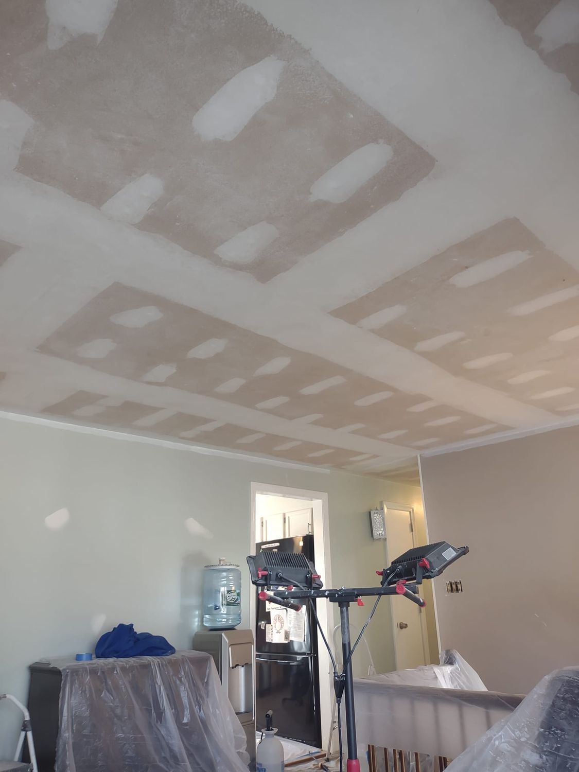 Living room under renovation with drywall patching on the ceiling and a treadmill near a water cooler