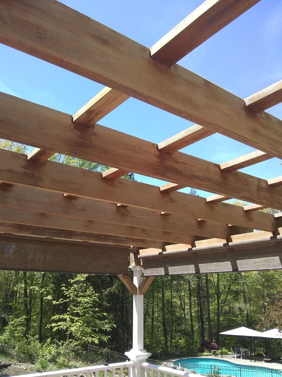 Wooden pergola beams over a backyard patio with a pool and umbrellas beyond