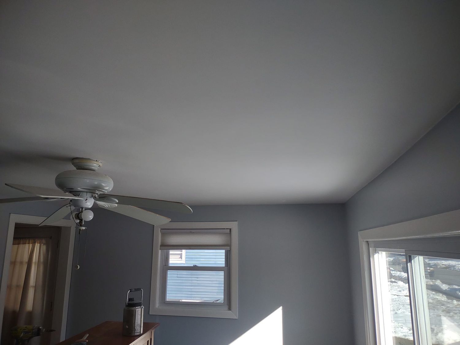 Empty gray bedroom with a ceiling fan, small windows, and sunlight on the wall.