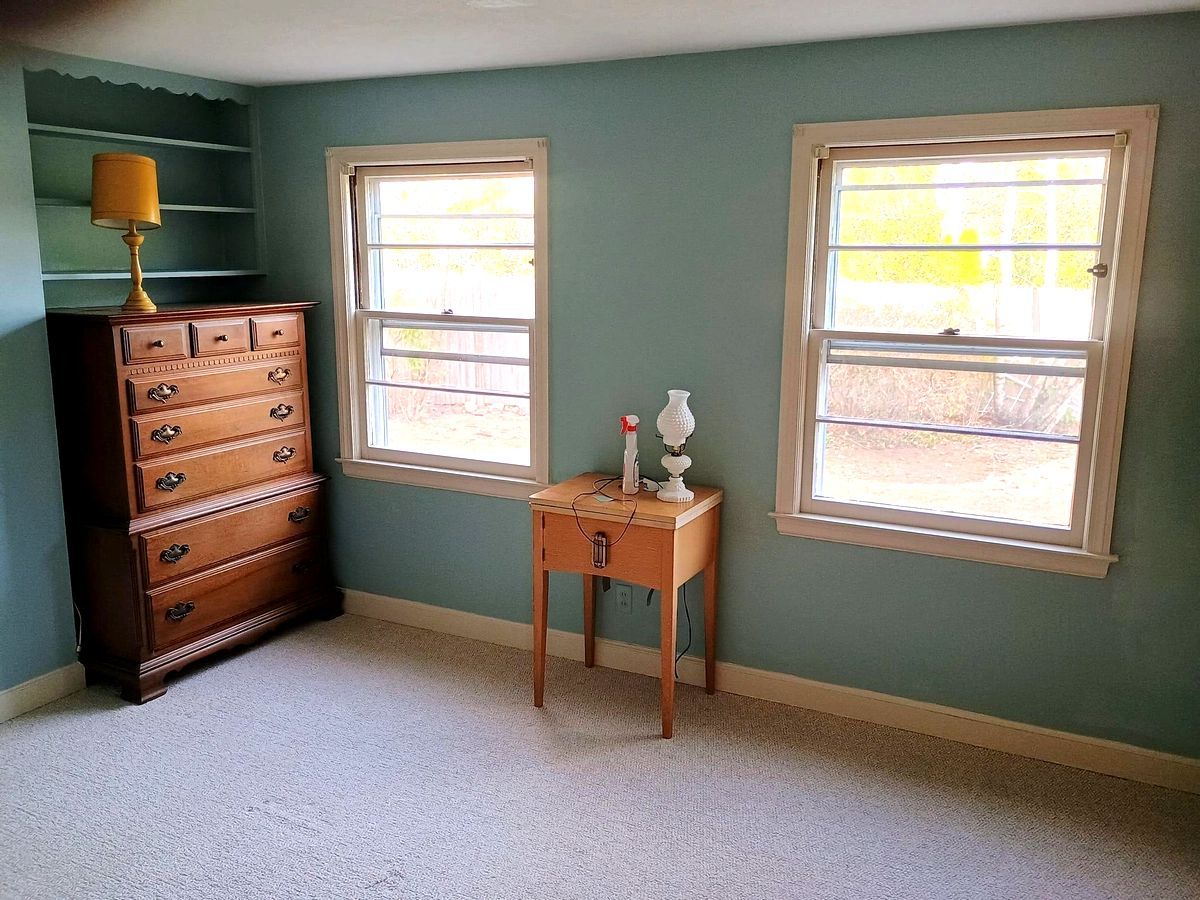 Empty bedroom with teal walls, two windows, a small table, and a wooden dresser with a lamp.