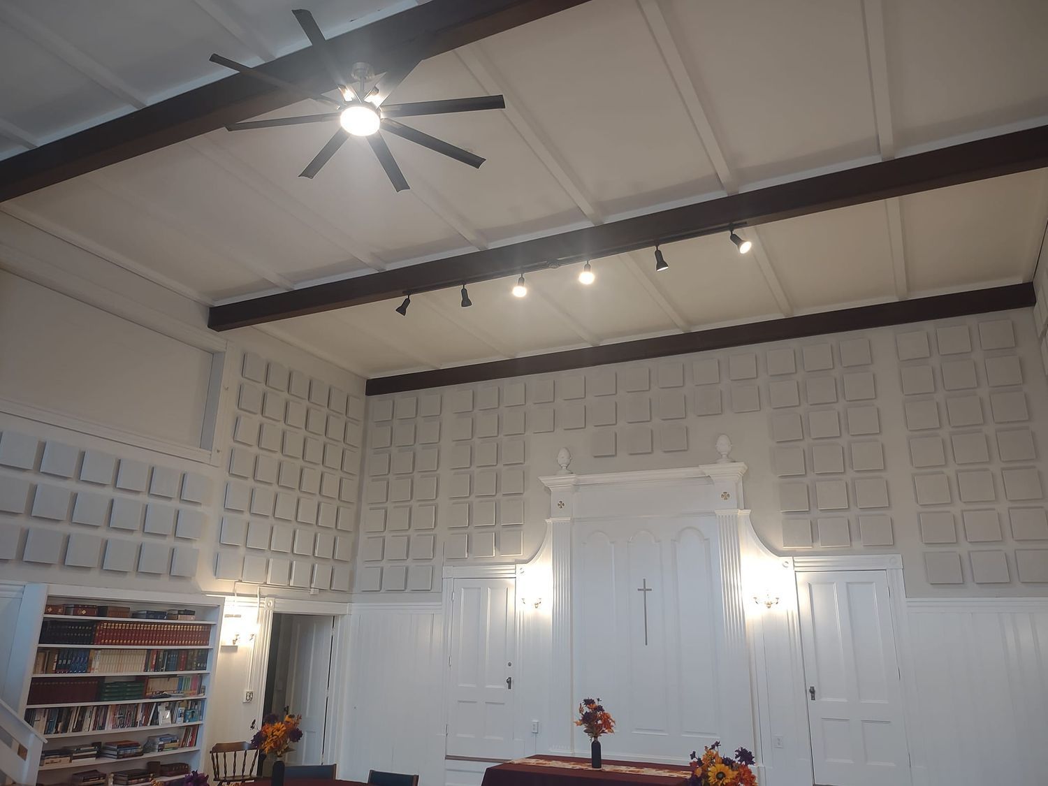 Interior of a room with a high ceiling, white paneled walls, ceiling fan, and lights above white double doors.