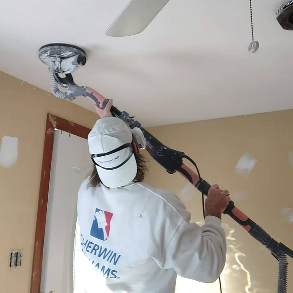 Painter sanding a ceiling corner in a room, wearing a Sherwin-Williams shirt and mask.
