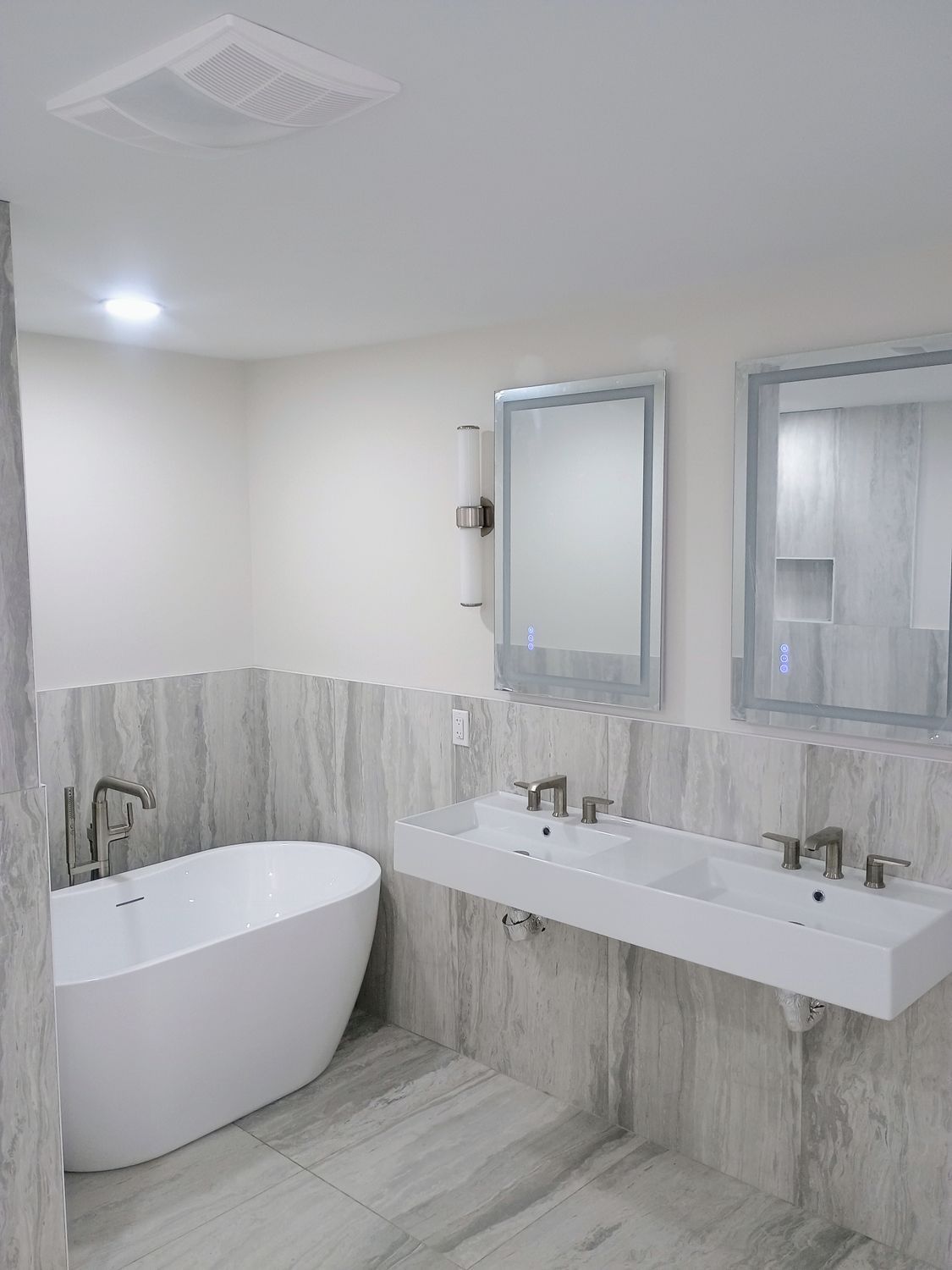 Modern bathroom with freestanding tub, double vanity, large mirrors, and gray tile walls and floor