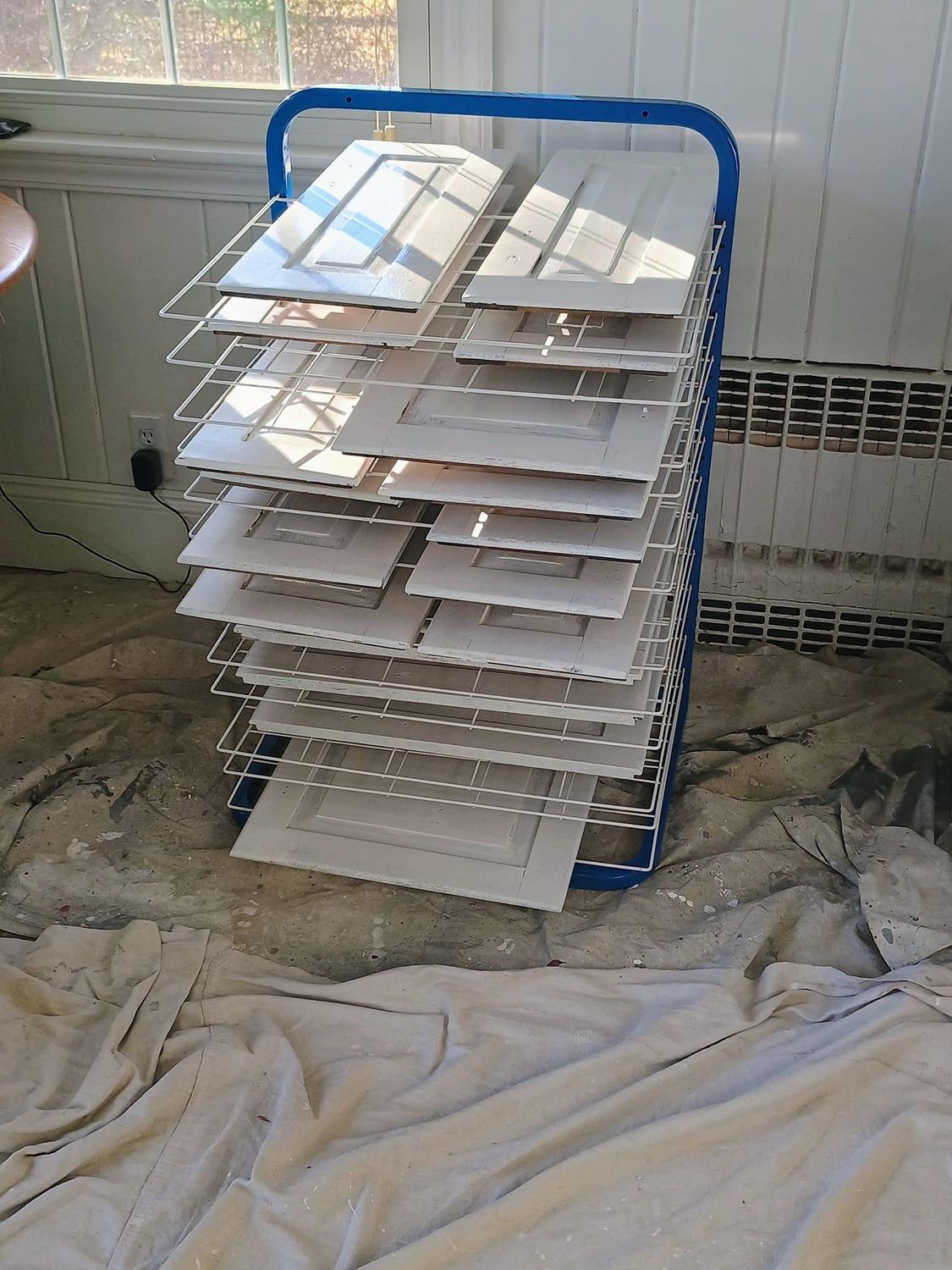 Blue wire rack holding stacked white trays in a room with a window and covered floor.