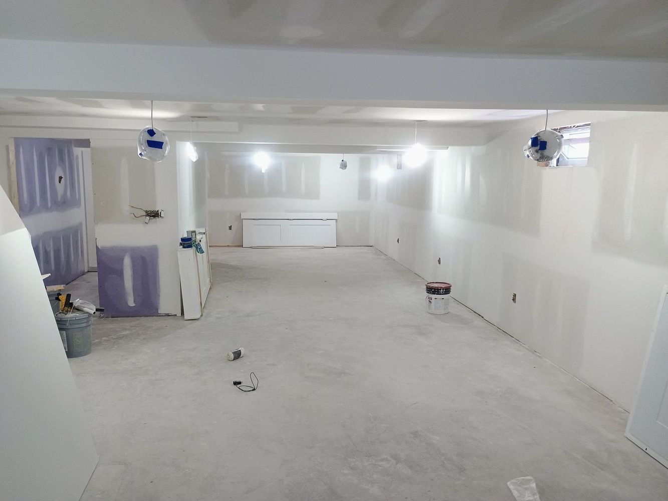 Empty white basement room under renovation with exposed ceiling lights and a work bucket on the floor