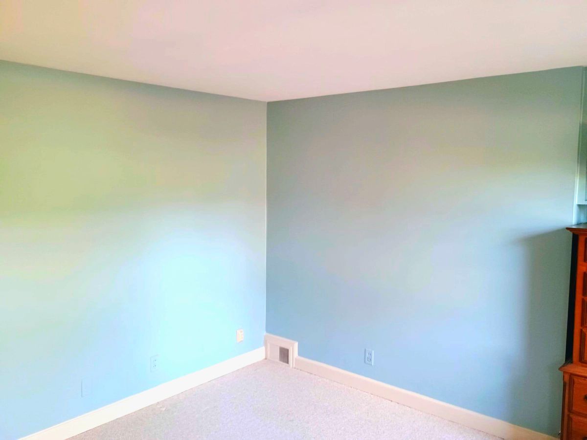 Empty light-blue room corner with white trim and carpet, partially showing a wooden dresser on the right