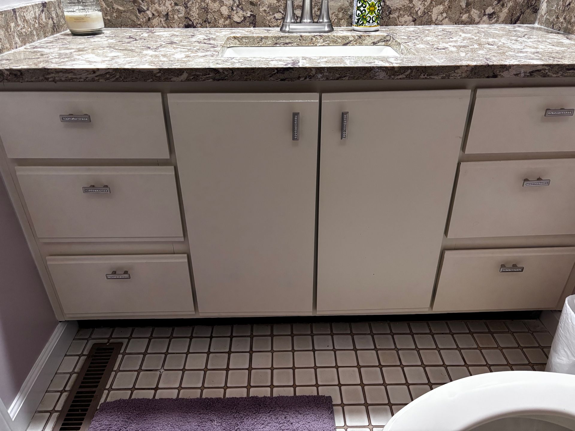 Bathroom vanity with beige cabinets, granite countertop, and white tiled floor with a purple bath mat
