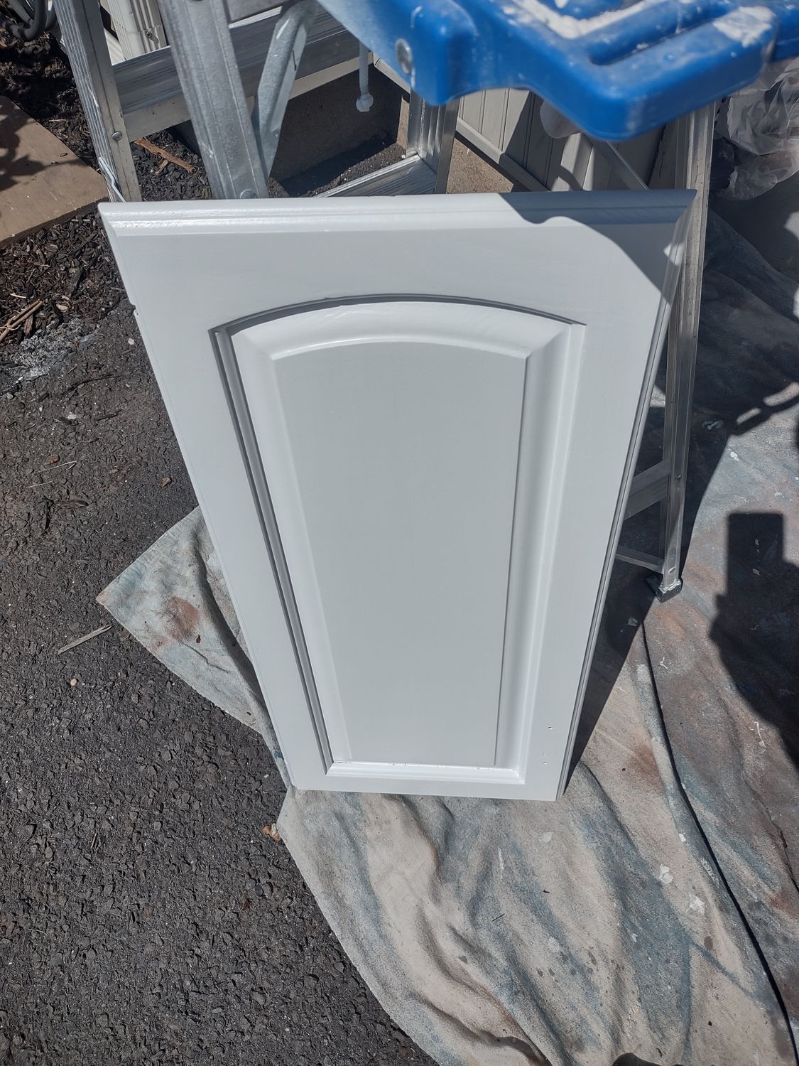 White cabinet door panel leaning outdoors on a tarp, with blue bins and gravel in the background