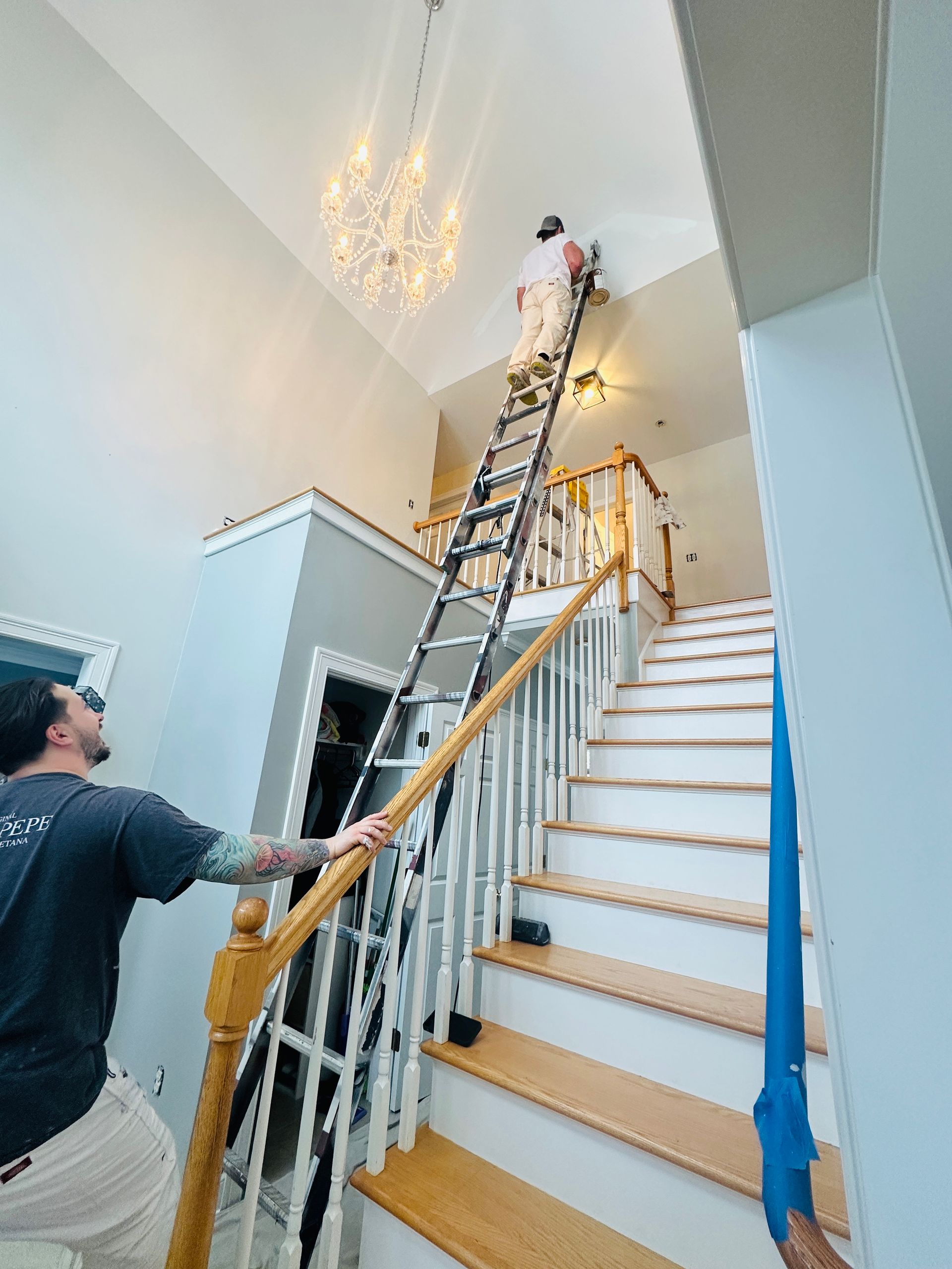 Two people on a ladder installing a chandelier in a bright stairwell with white walls and wooden steps