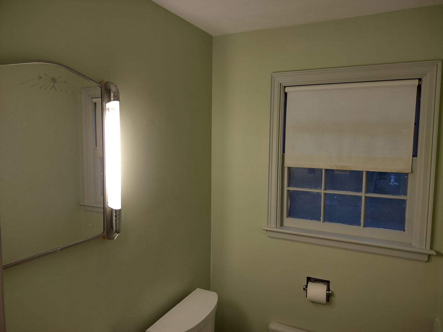 Small bathroom with green walls, mirror, window with blind, and towel ring above the toilet paper holder