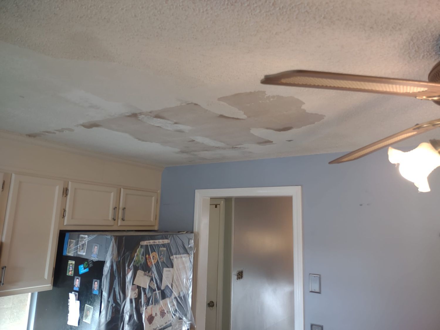 Kitchen ceiling with water damage and peeling paint above white cabinets and a stainless refrigerator