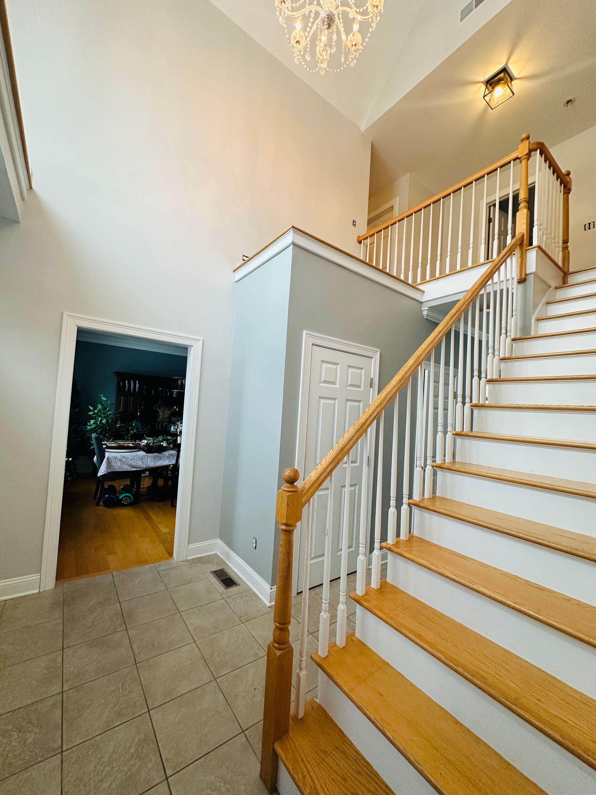 Bright foyer with white staircase, wooden railings, and beige tiled floor, viewed from the bottom stairs