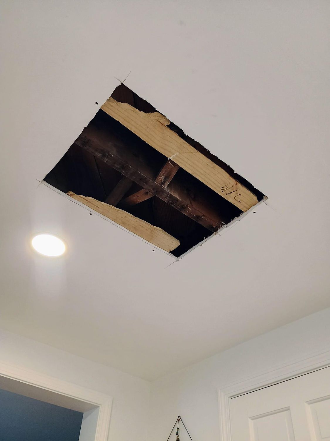 Ceiling with a rectangular hole exposing wooden framing and a light fixture nearby
