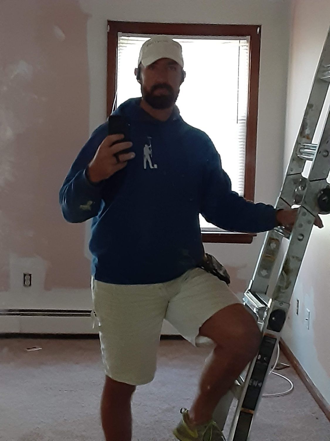 Man posing indoors on a ladder in a room under renovation, wearing a blue hoodie and khaki shorts.