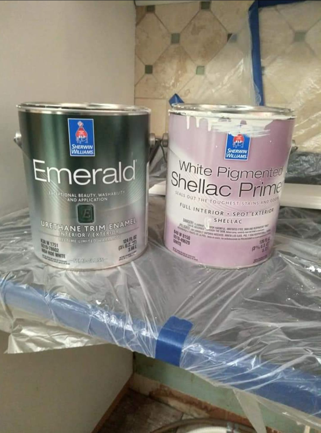 Two paint cans on a wrapped shelf, one green Emerald and one pink White Pigeon shellac primer.
