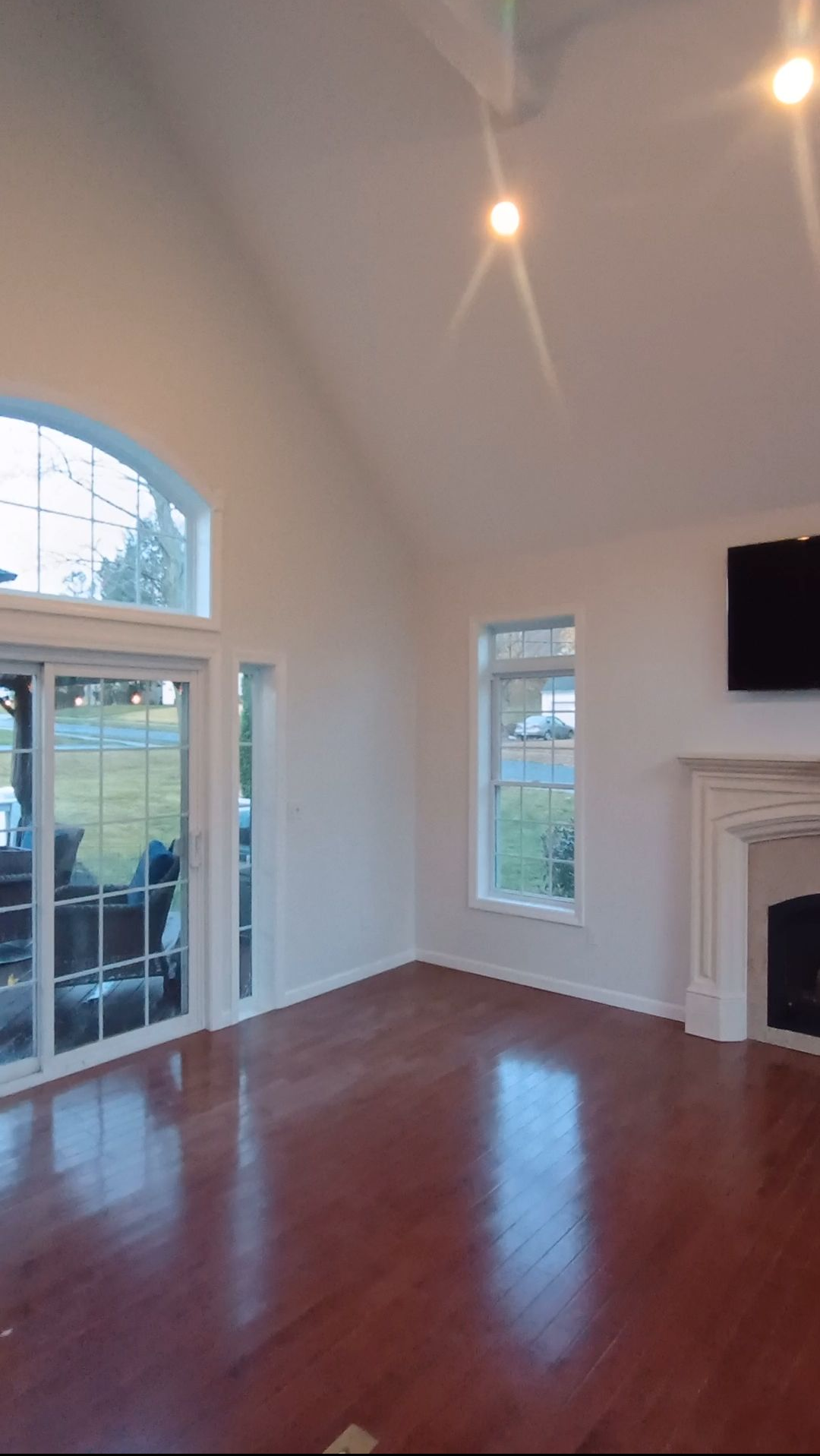 Empty living room with hardwood floors, tall arched windows, and a fireplace