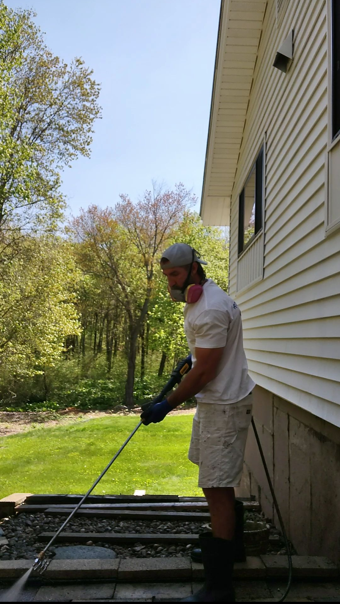 Person using a long tool beside a house, working on a raised deck in a sunny yard