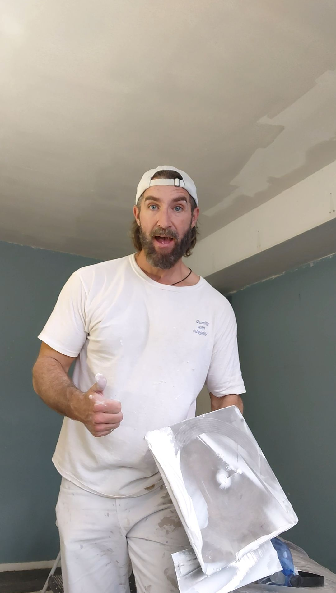Man in white shirt and backward cap holding a paint tray and giving a thumbs-up in a room under renovation