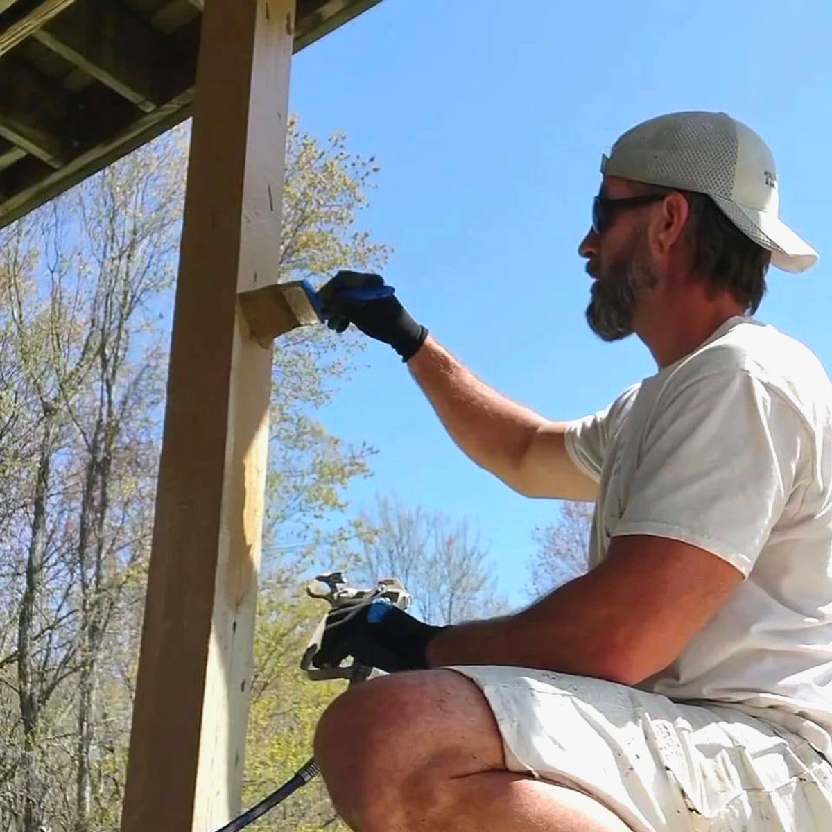 Worker painting a porch post outdoors in sunlight, wearing gloves and a cap