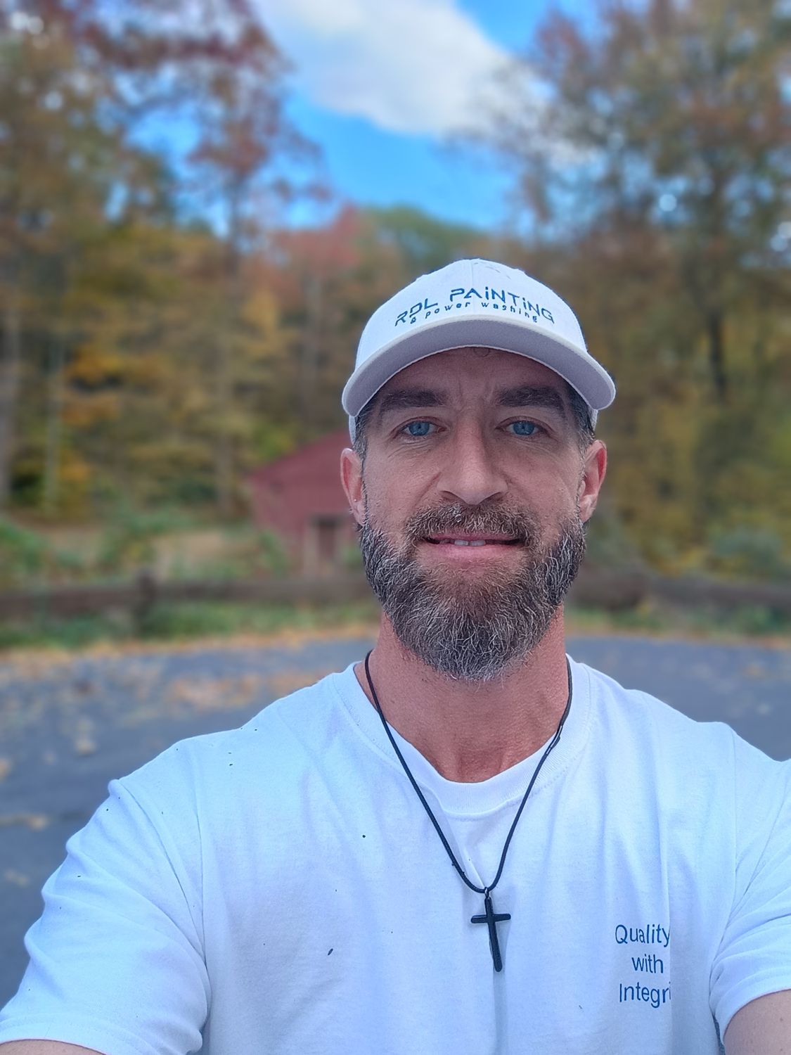 Man smiling in a white cap and shirt, taking a selfie outdoors with autumn trees and a blue sky.
