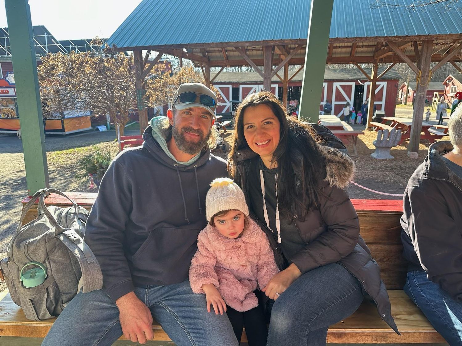 Family of three sitting on a bench under a pavilion, smiling outdoors on a sunny day.