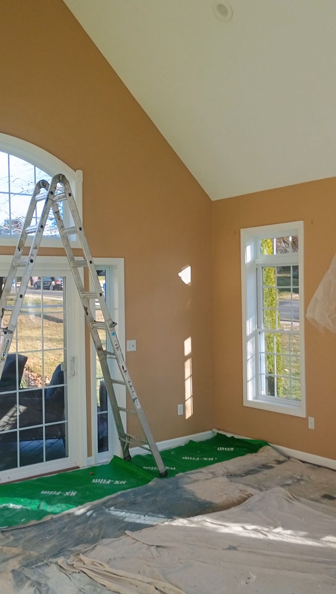 Interior room under renovation with orange walls, tall windows, a ladder, and floor protection sheets