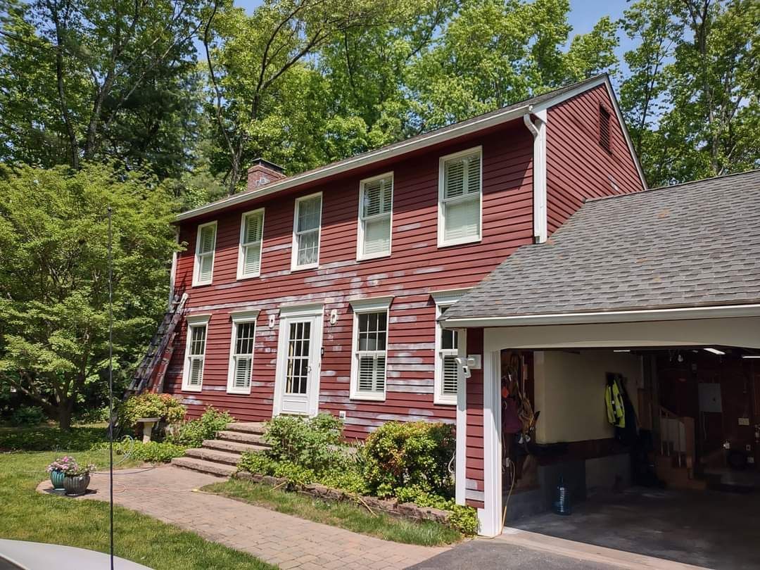 Red two-story house with white trim and attached carport, surrounded by trees and lawn