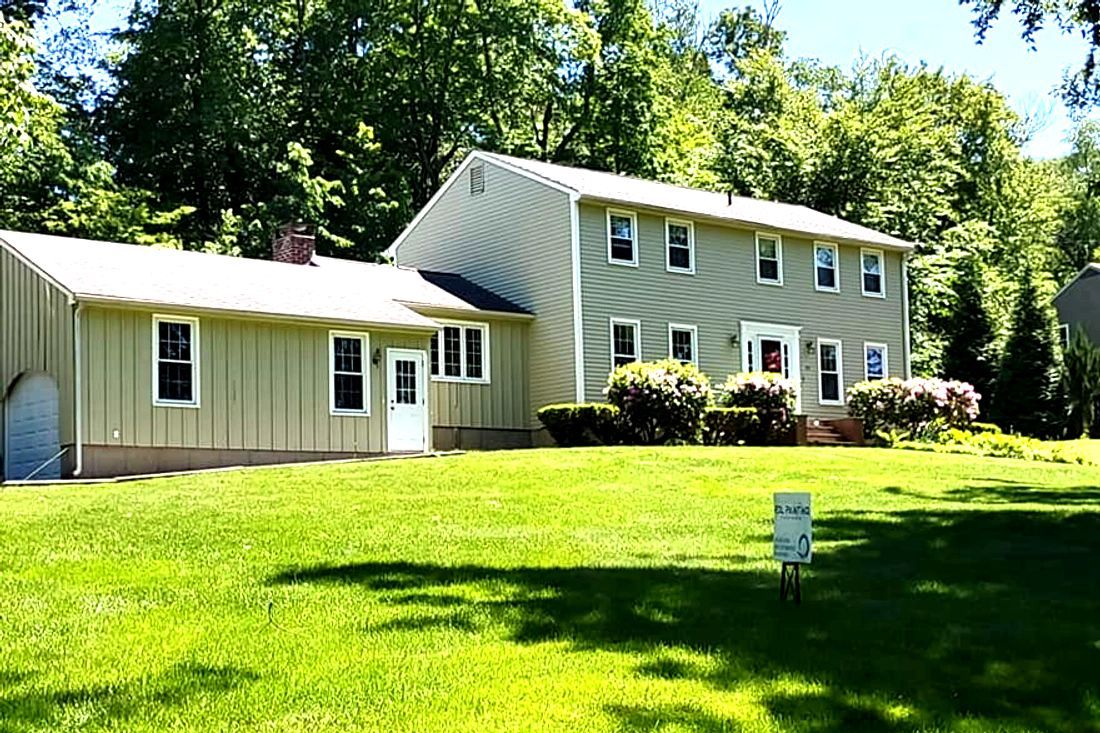A beige house and attached garage in a sunny green yard with trees behind it.