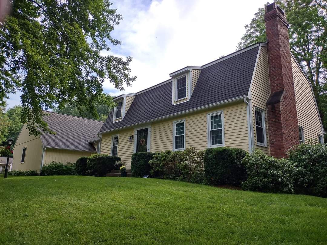 Yellow house with dark roof, white trim, and brick chimney beside a green lawn and trees