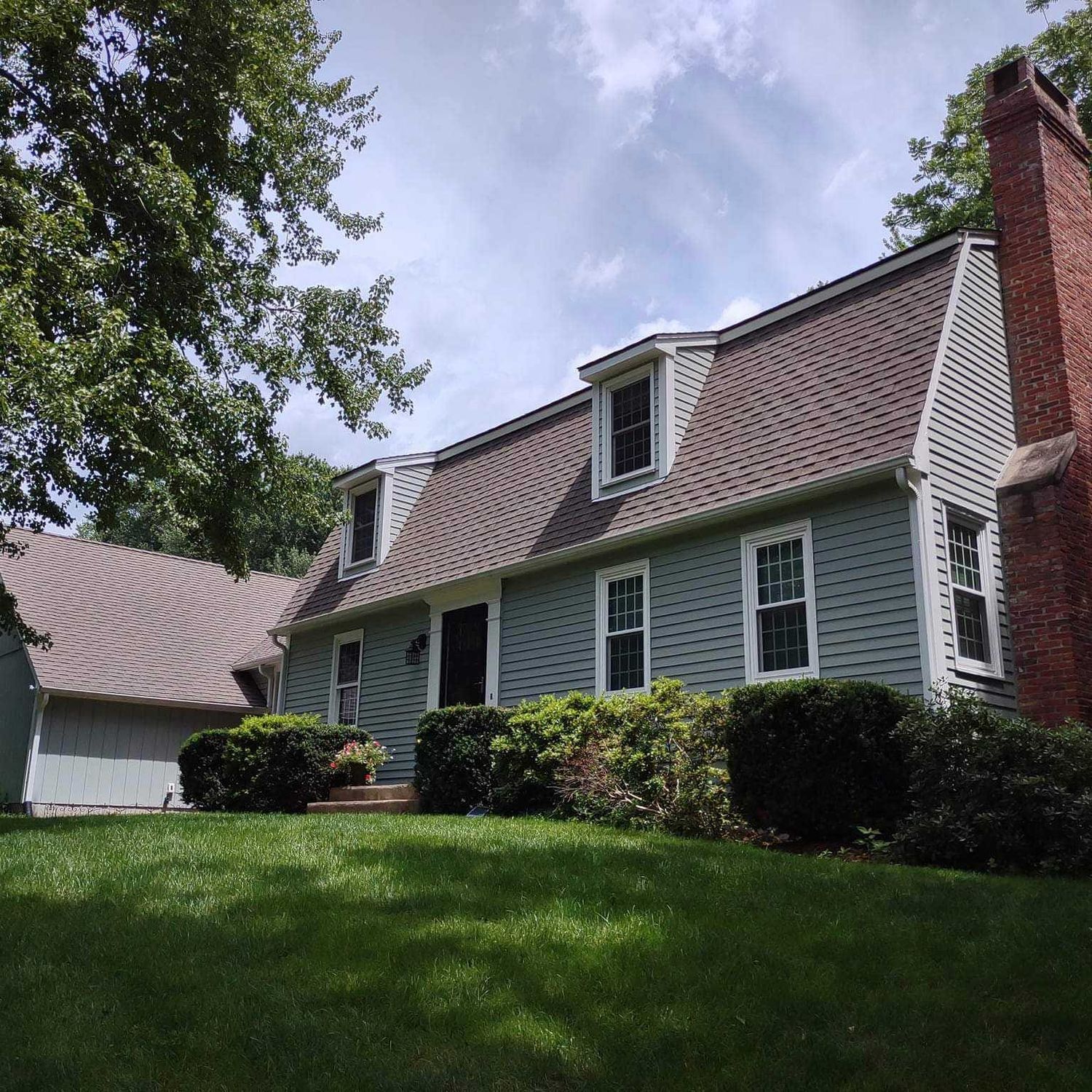 White house with red shingled roof, dormer windows, and brick chimney behind a green lawn and trees.