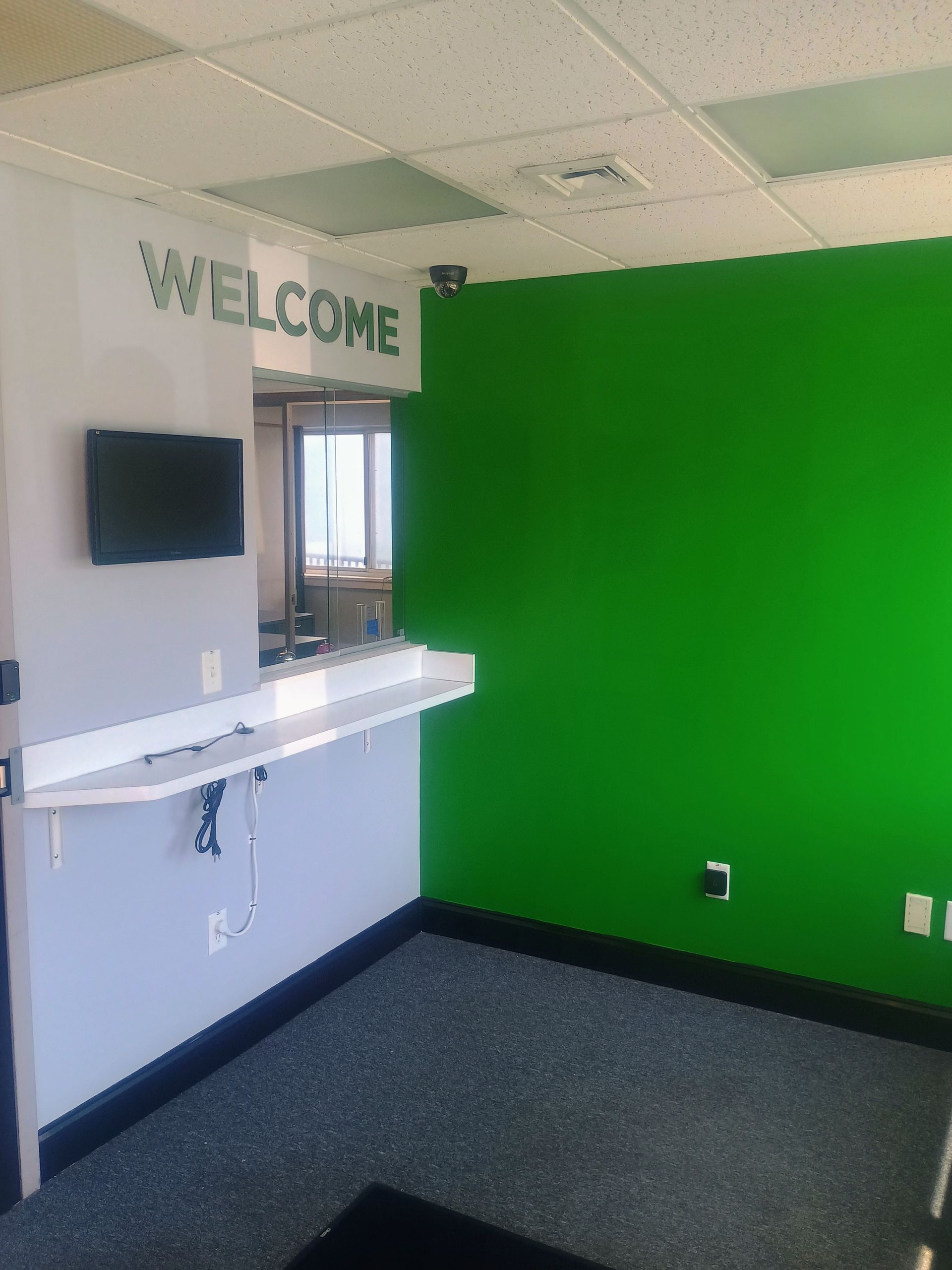 Small lobby with a green accent wall, white counter, wall-mounted screen, and “WELCOME” sign.