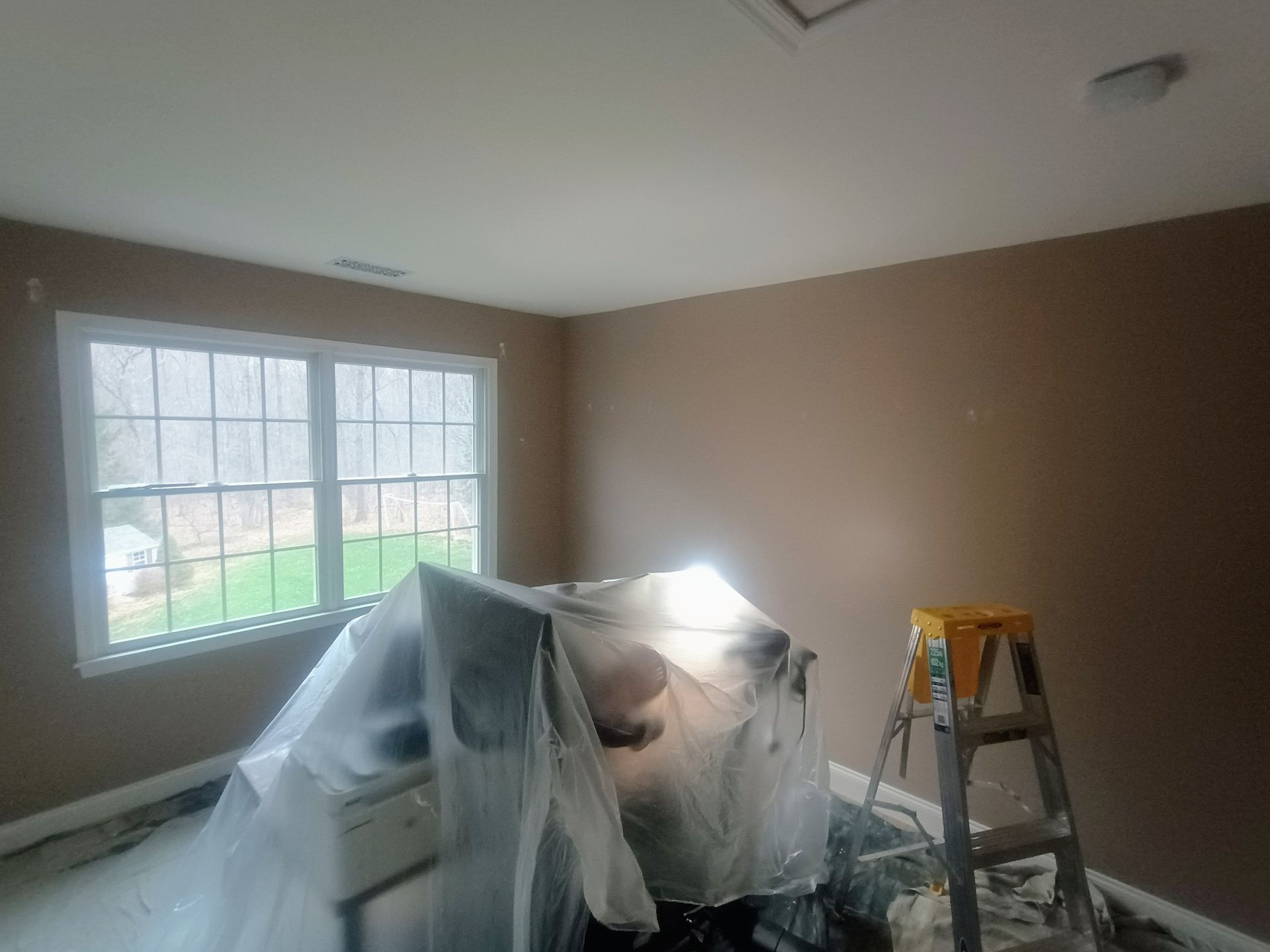 Room under renovation with plastic-covered furniture, ladder, and sunlight through a large window