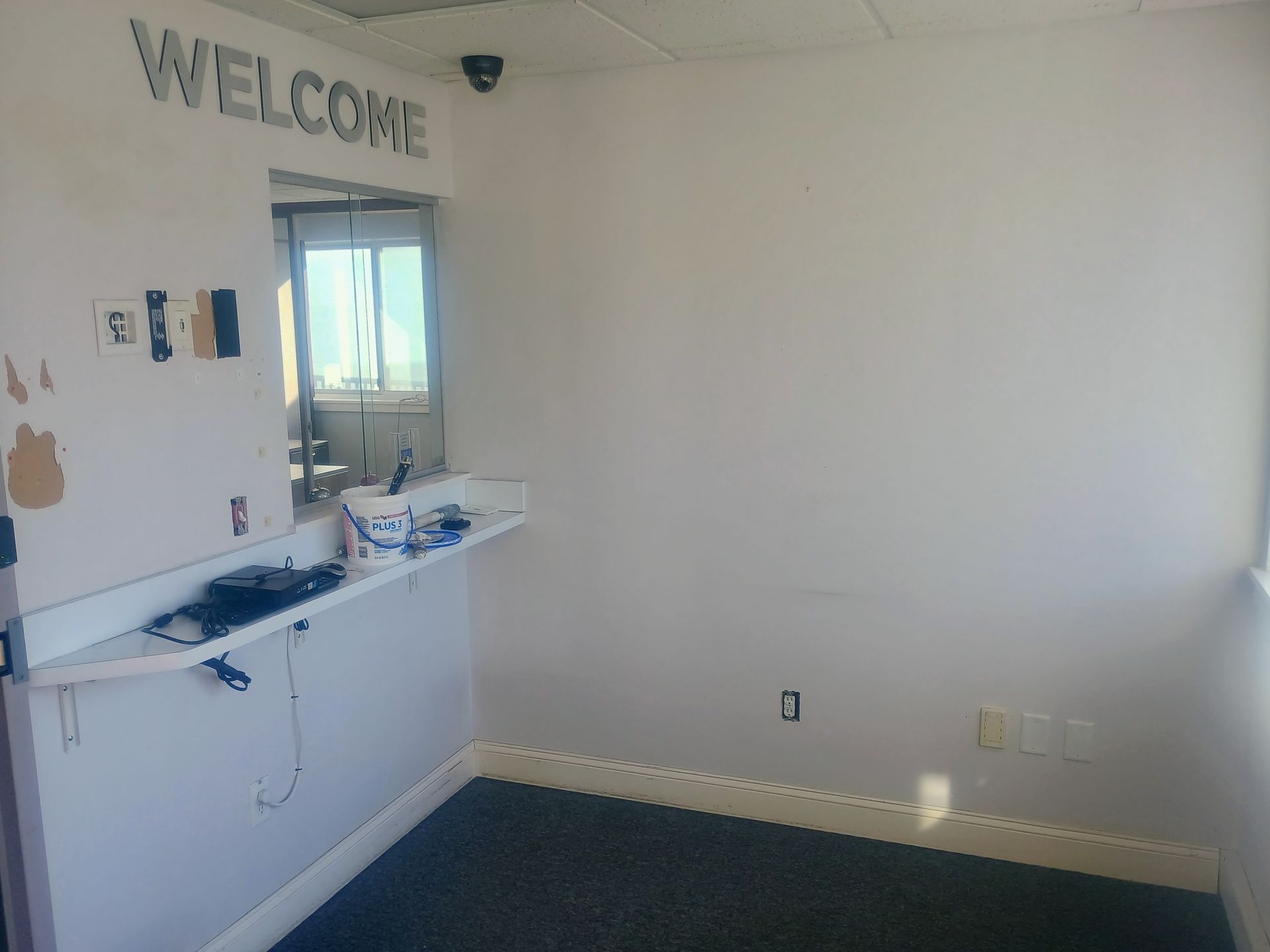 Small white reception room with a counter, mirror, “WELCOME” sign, and dark carpeted floor.