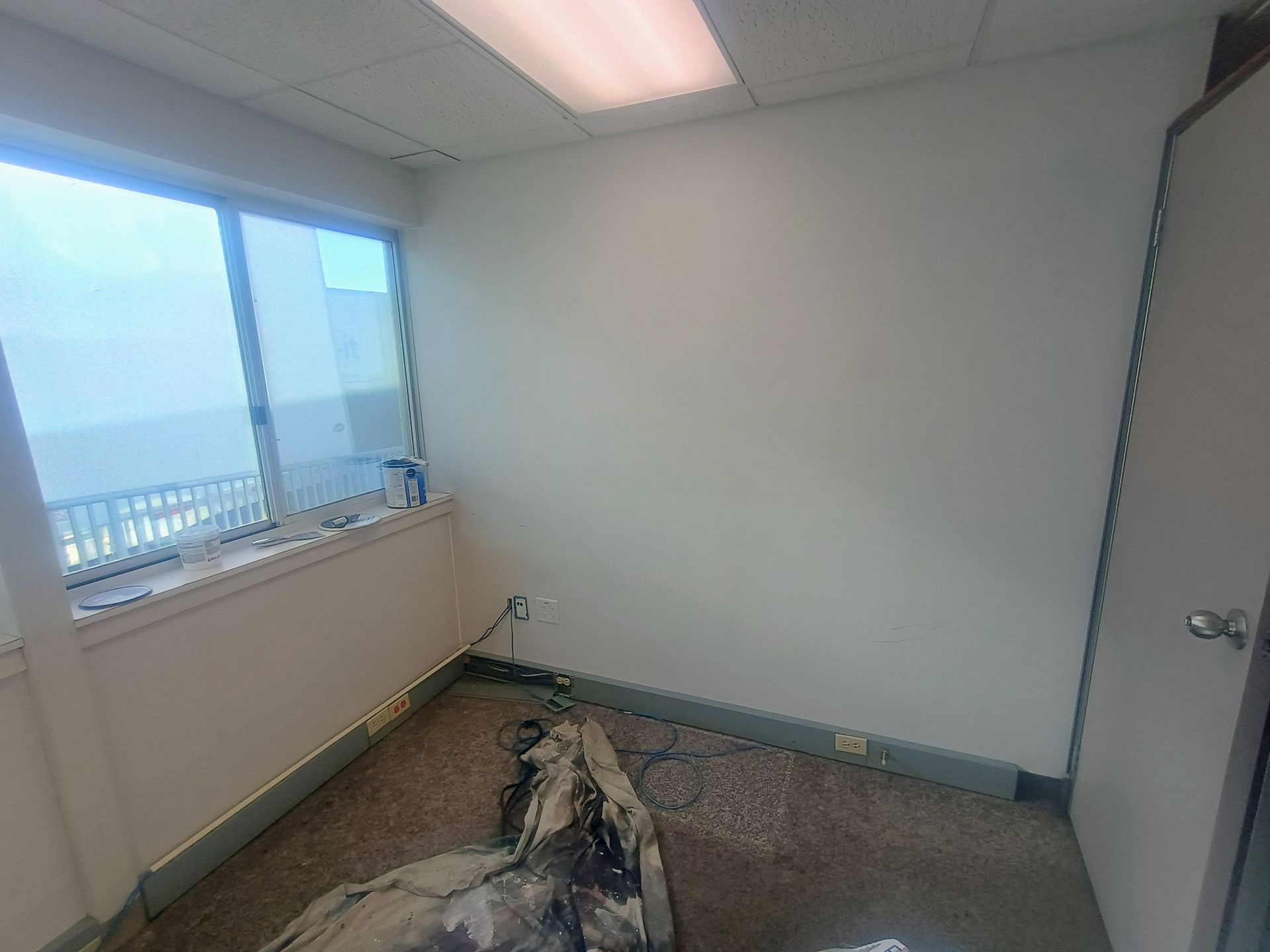 Empty office corner with damaged carpet, a window on the left, and a closed door on the right.