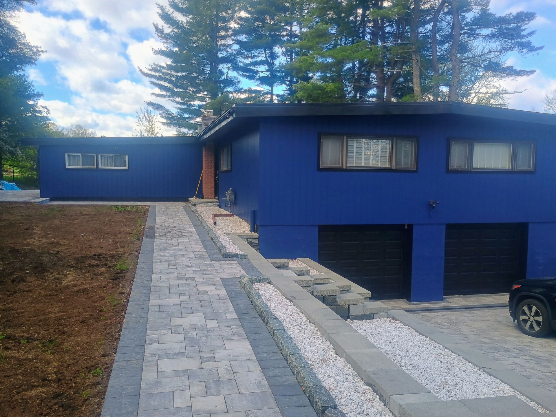 Blue house with a stone walkway and driveway, backed by tall trees and a partly cloudy sky