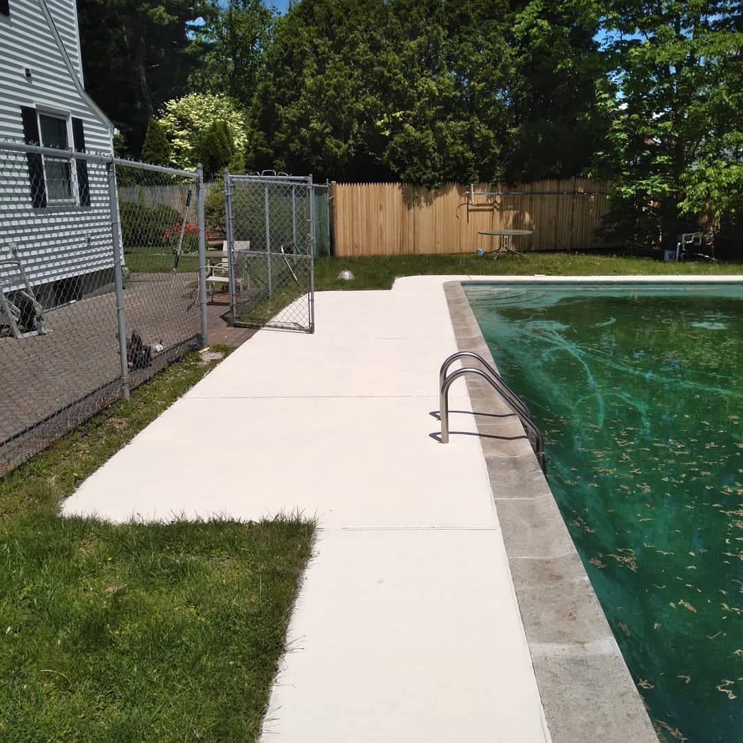 Backyard pool with white concrete walkway, metal handrail, and fenced lawn beside a house.